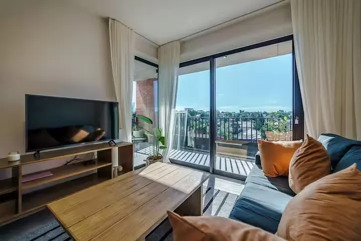 Cozy sunlit living room in off-season vacation rental, with sofa, TV, coffee table and balcony overlooking quiet rooftops.