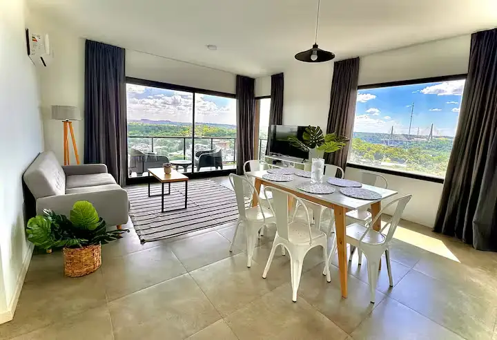 Bright riverside vacation apartment living and dining area with balcony views of a quiet river and bridge in the off-season.