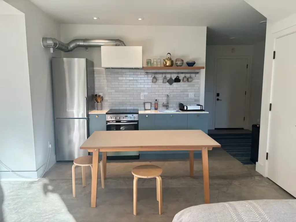 Empty minimalist kitchenette in a quiet off-season rental with sunlit table, stools, concrete floor and tile backsplash.