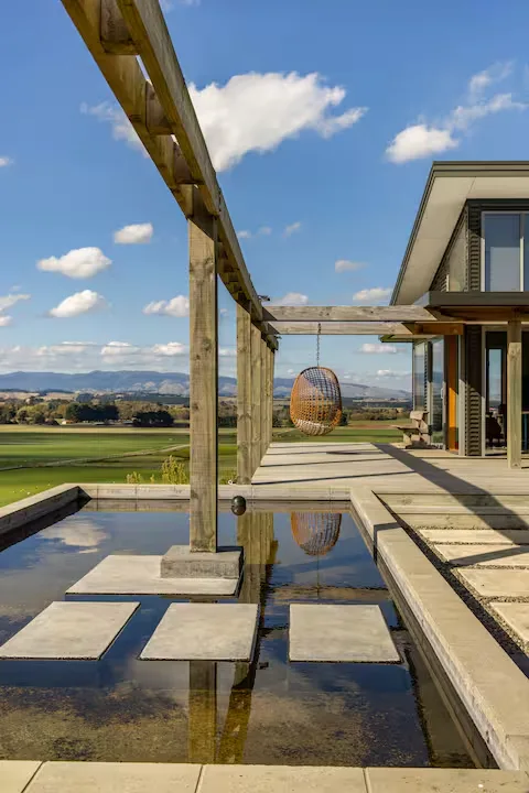 Serene off-season retreat with modern terrace, hanging wicker chair over reflecting pool and green fields under clear sky.