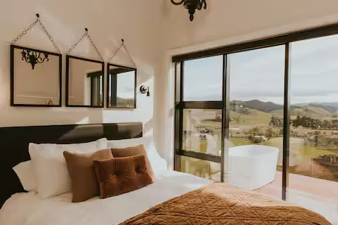 Sunlit hotel room with cozy bed and pillows opening onto a quiet balcony view of empty rolling green hills in off-season.
