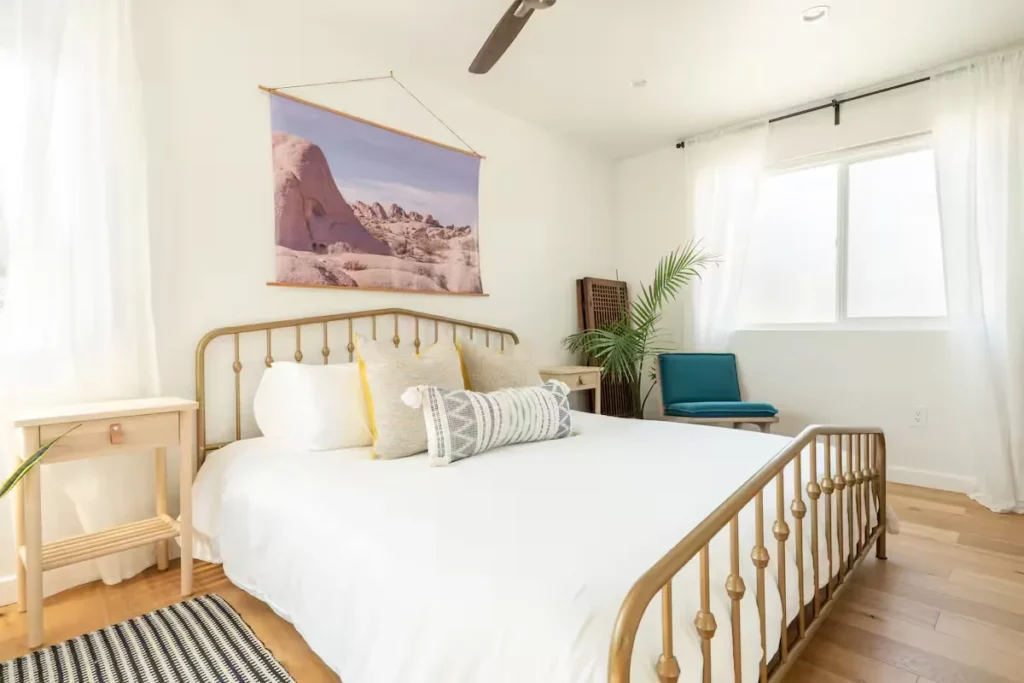 Airy off-season vacation rental bedroom with soft natural light, gold metal bedframe, white linens and desert landscape art.