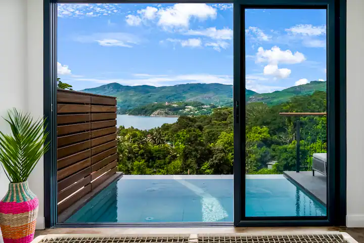 Infinity pool through sliding glass doors overlooking lush hills and a calm seaside bay in off-season serenity.