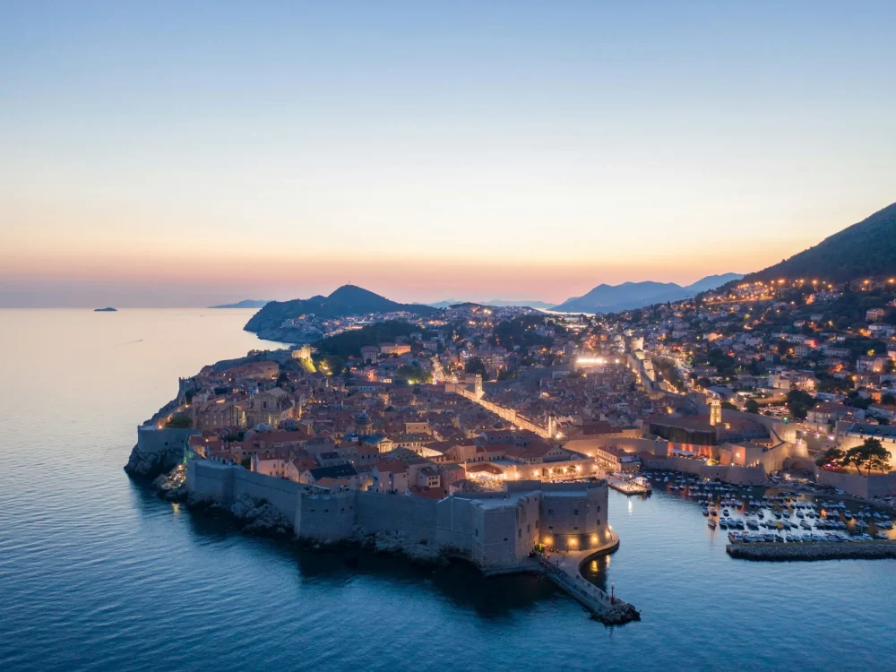 An aerial view of the walled city of Dubrovnik at dusk, with lights twinkling along the coastline.